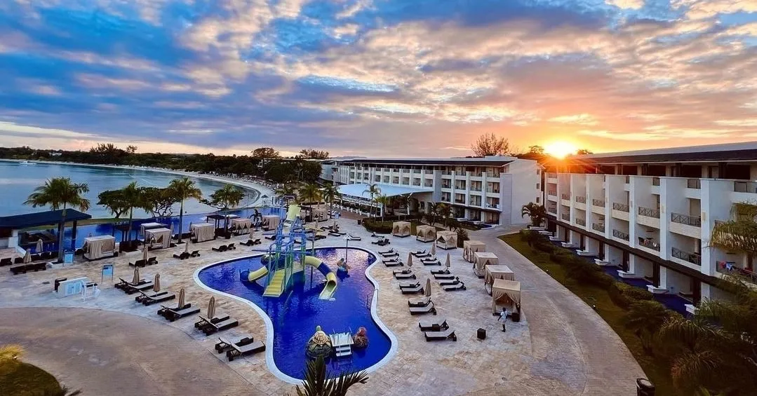 A beach resort at sunset, its pool and cabanas in the foreground and a calm curve of bay beyond.