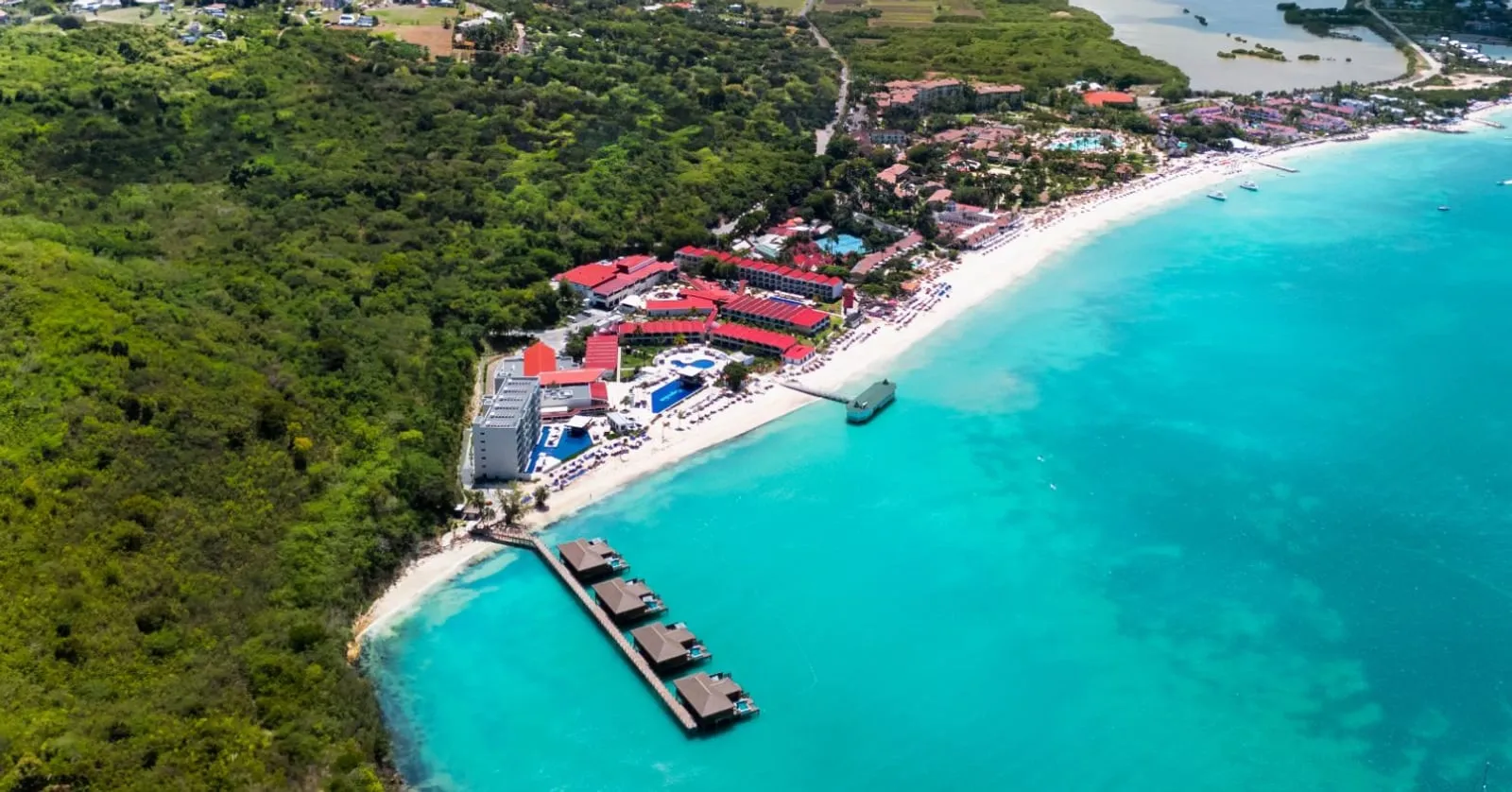 Aerial view of a beachfront resort on a curve of white sand, with overwater villas on a pier over turquoise shallows.
